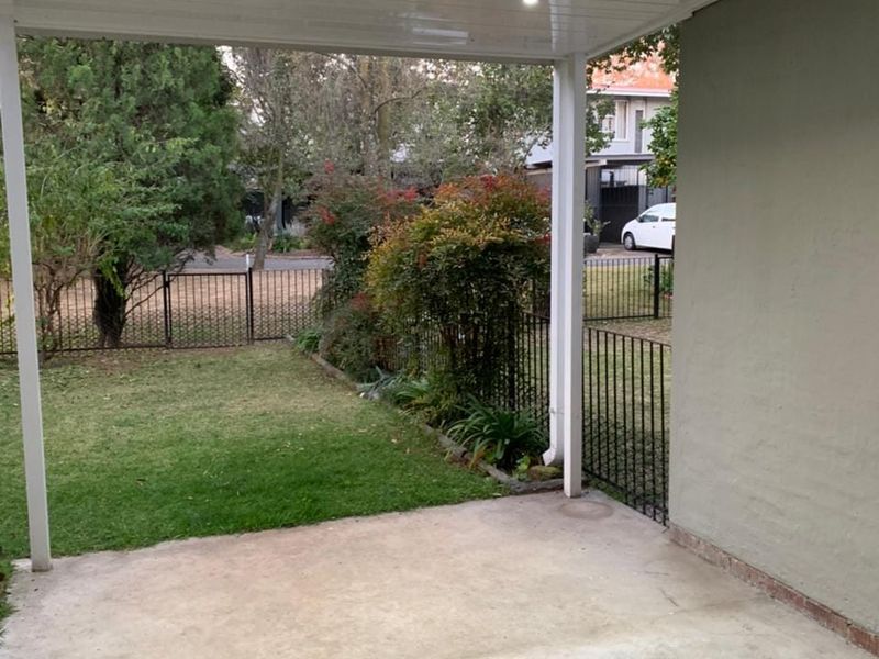 A covered patio area featuring concrete flooring and recessed ceiling lights, overlooking a private garden with metal perimeter fencing.