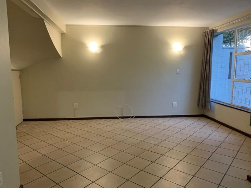 An empty living room featuring neutral tiled flooring, wall-mounted lighting, and a window with security bars, offering a blank canvas for interior design.