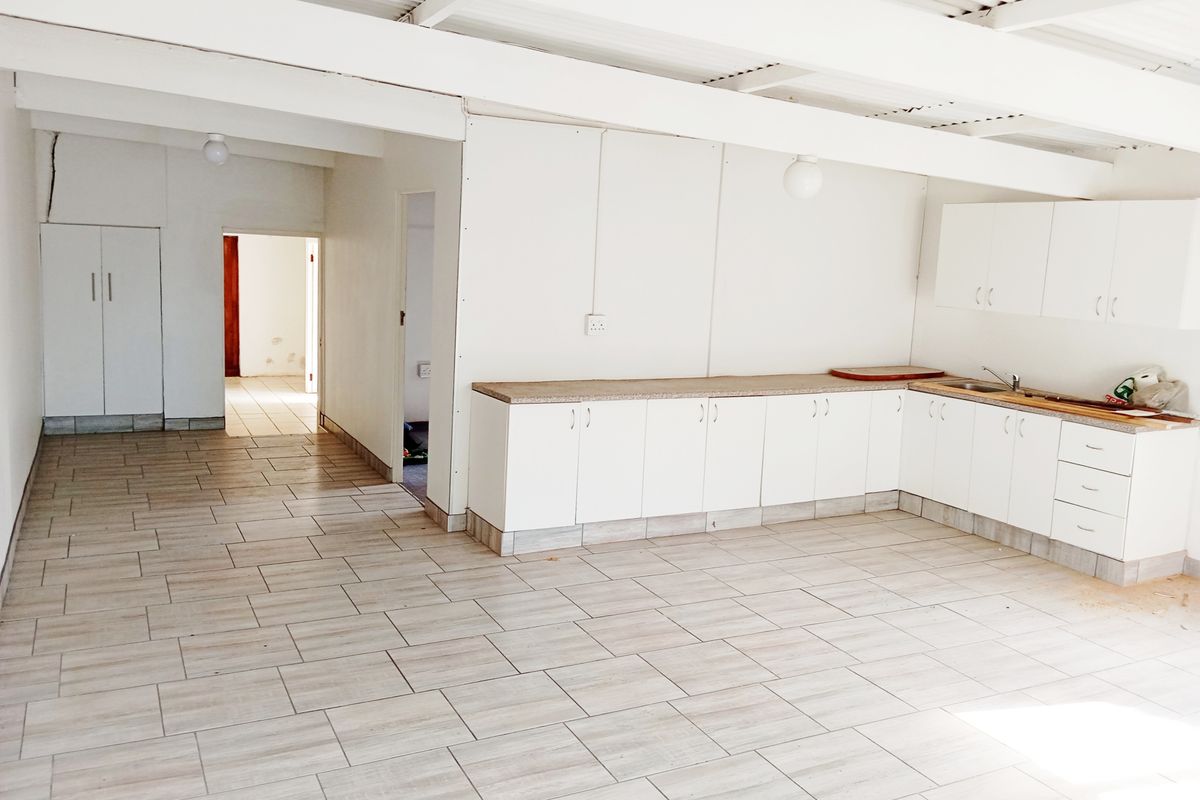 An empty kitchen area featuring white built-in cabinetry, laminate countertops, a stainless steel sink, and tiled flooring under an exposed beam ceiling.