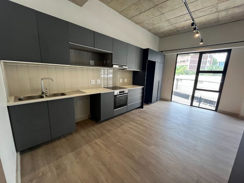 Modern open-plan kitchen featuring dark cabinetry, integrated appliances, and exposed concrete ceilings, flowing into a bright living area with large glass doors.