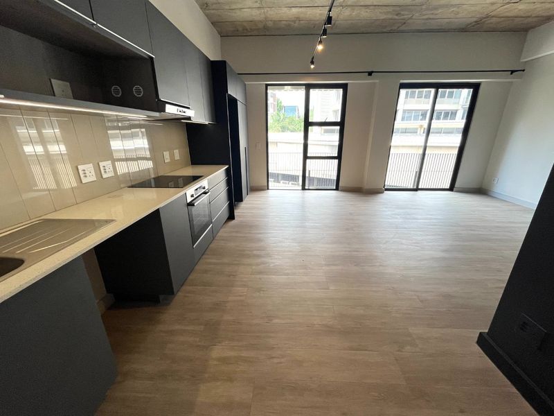 Modern open-plan kitchen and living area featuring sleek dark cabinetry, stone countertops, exposed concrete ceilings, and large sliding doors leading to a balcony.