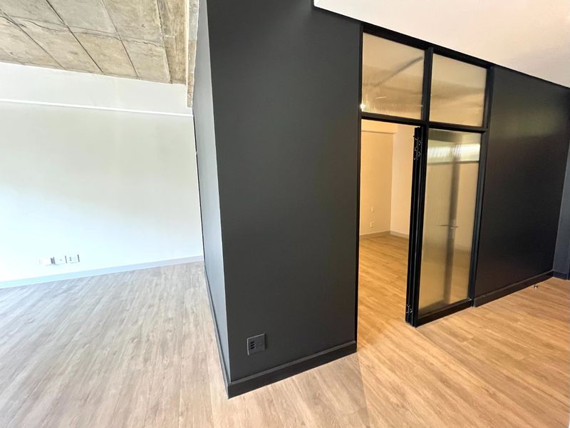 Modern open-plan living area featuring exposed concrete ceilings, light wood-look laminate flooring, and a stylish black-framed glass partition door.