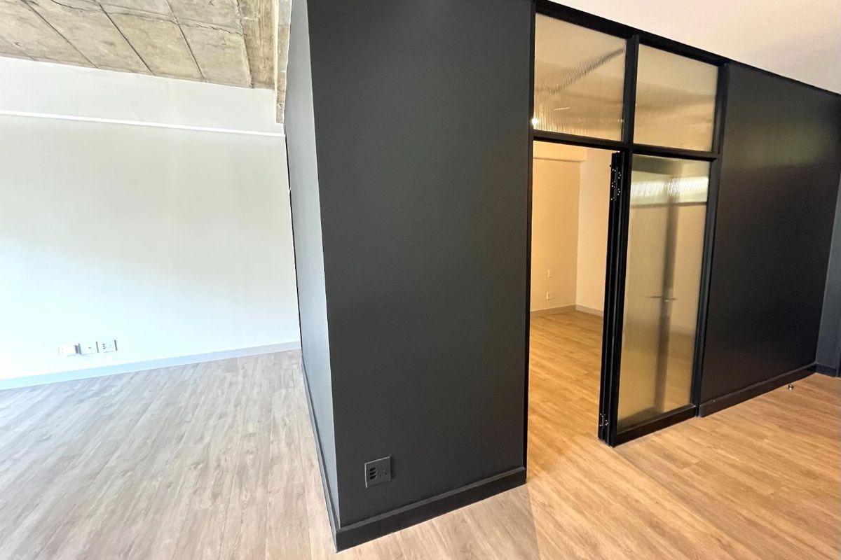 Modern open-plan living area featuring exposed concrete ceilings, light wood-look laminate flooring, and a stylish black-framed glass partition door.