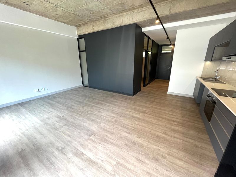 Modern open-plan living area featuring exposed concrete ceilings, sleek dark cabinetry, integrated kitchen appliances, and durable laminate flooring in a contemporary apartment setting.
