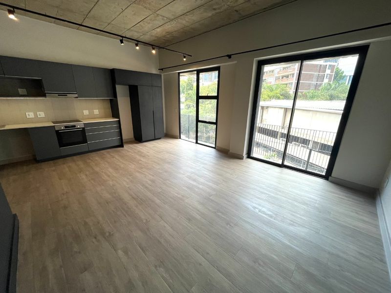 Modern open-plan living area featuring an exposed concrete ceiling, sleek dark cabinetry, integrated kitchen appliances, and large sliding doors leading to a balcony.