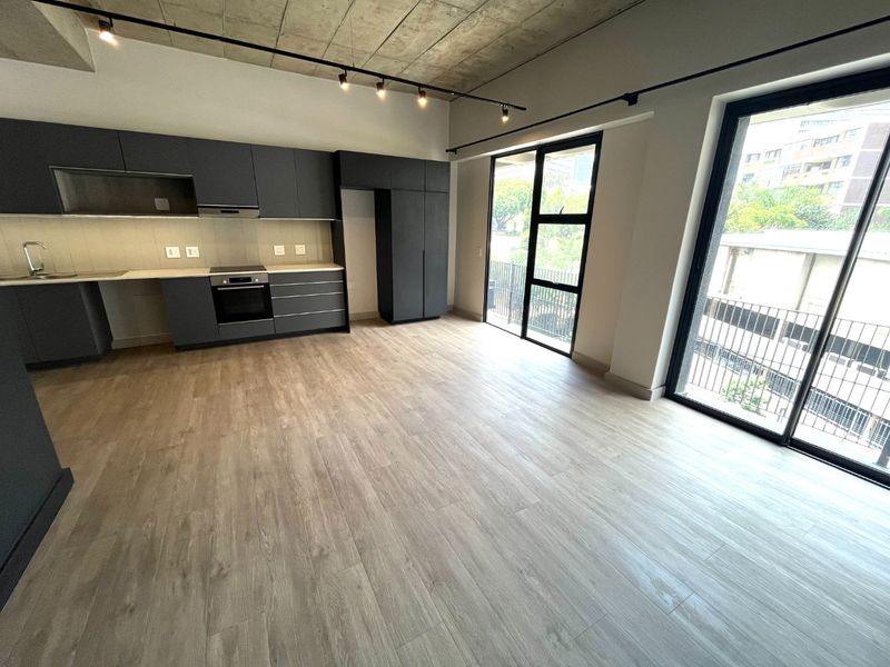 Modern open-plan kitchen and living area featuring sleek dark cabinetry, exposed concrete ceilings, track lighting, and large sliding doors leading to a balcony.