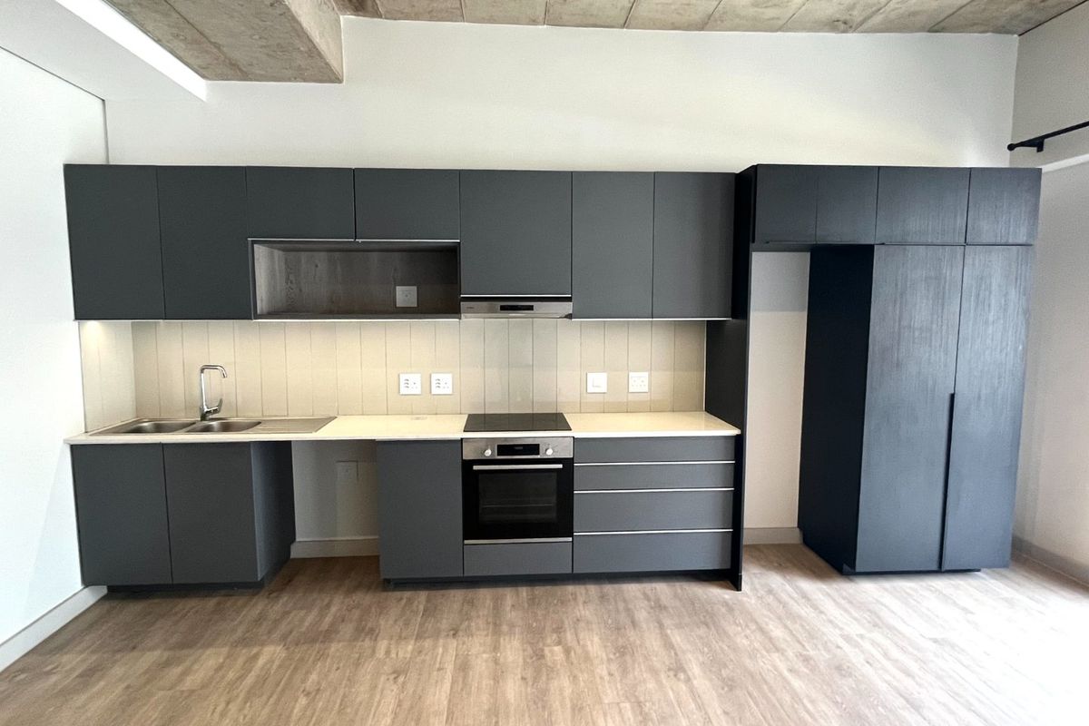 Modern kitchen featuring dark grey cabinetry, stone countertops, integrated oven and hob, and an exposed concrete ceiling in an open-plan layout.