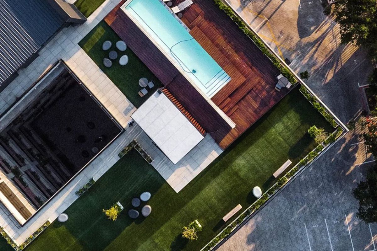 Aerial view of a modern apartment complex featuring a swimming pool with timber decking, landscaped green spaces, and paved parking facilities.