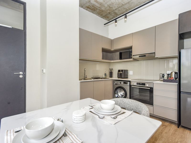 Modern open-plan kitchen featuring sleek cabinetry, integrated appliances, track lighting, and an exposed concrete ceiling, seamlessly flowing into the dining area.