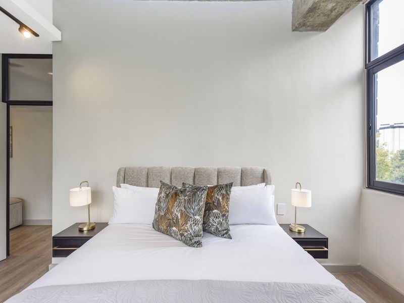 A modern bedroom featuring laminate flooring, an exposed concrete beam, and a large window providing natural light.
