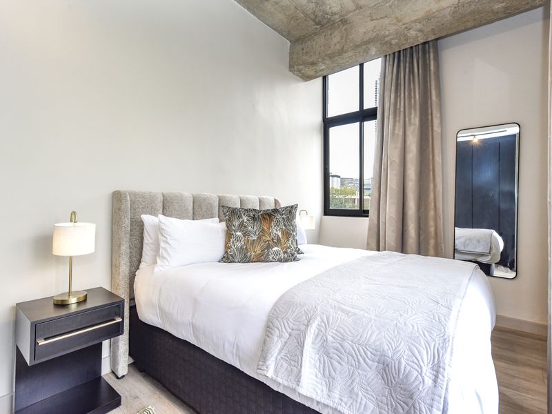A modern bedroom featuring an exposed concrete ceiling beam, large black-framed window, and light laminate flooring, creating a contemporary industrial aesthetic.