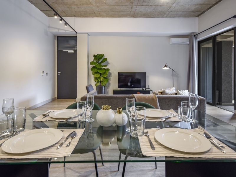 Modern open-plan living and dining area featuring an exposed concrete ceiling, track lighting, and seamless access to an outdoor patio through sliding glass doors.