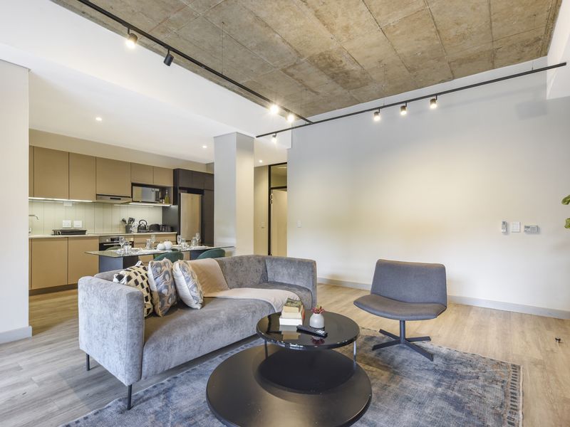 Modern open-plan living area featuring an exposed concrete ceiling, track lighting, and a sleek, contemporary kitchen with integrated appliances and laminate flooring.
