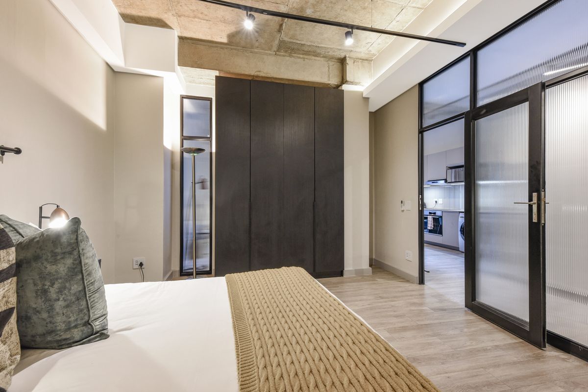 Modern bedroom featuring an exposed concrete ceiling, built-in dark cabinetry, and a stylish fluted glass door leading to the kitchen area.