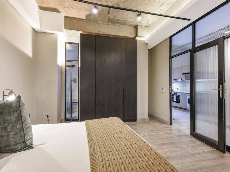 Modern bedroom featuring an exposed concrete ceiling, built-in dark cabinetry, and a stylish fluted glass door leading to the kitchen area.