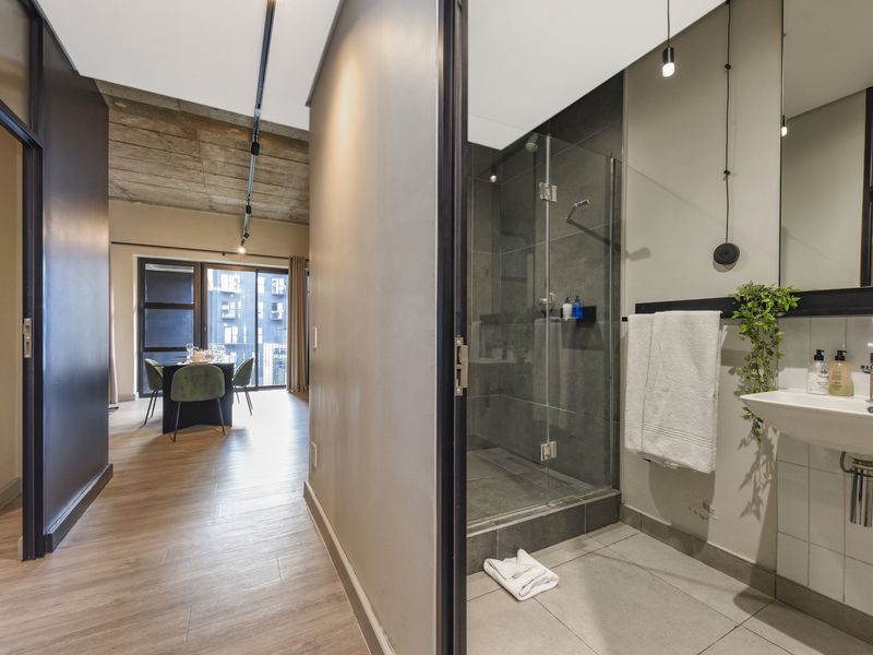 Modern bathroom featuring a glass-enclosed shower and wall-mounted basin, adjacent to an open-plan living area with exposed concrete ceilings.
