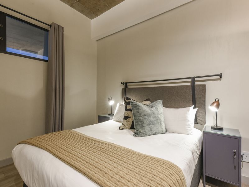 A modern bedroom featuring an exposed concrete ceiling, laminate flooring, and a high-level window, creating a contemporary industrial aesthetic.