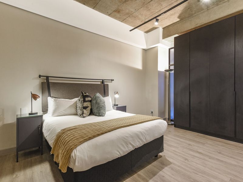 Modern bedroom featuring an exposed concrete ceiling, track lighting, laminate flooring, and floor-to-ceiling built-in cupboards.