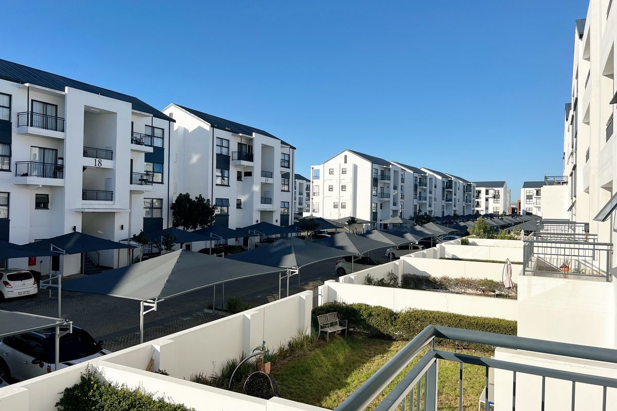 A view of the apartment complex exterior, featuring modern white building facades, paved parking areas with shade cloth carports, and landscaped garden sections.