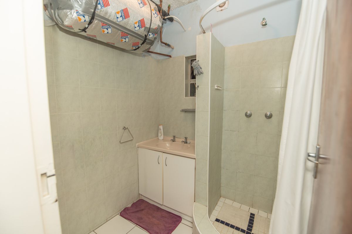 A dated bathroom featuring a vanity basin and shower area with exposed geyser plumbing and visible wear on wall and floor tiles.