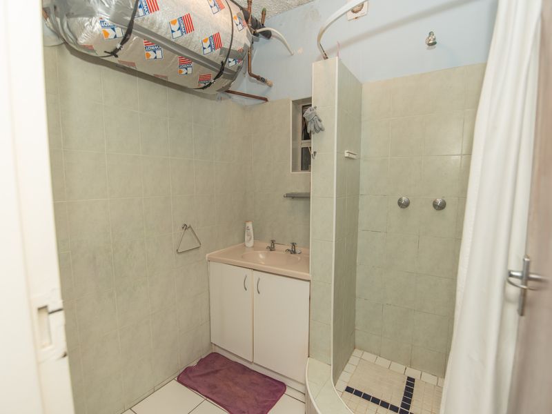 A dated bathroom featuring a vanity basin and shower area with exposed geyser plumbing and visible wear on wall and floor tiles.