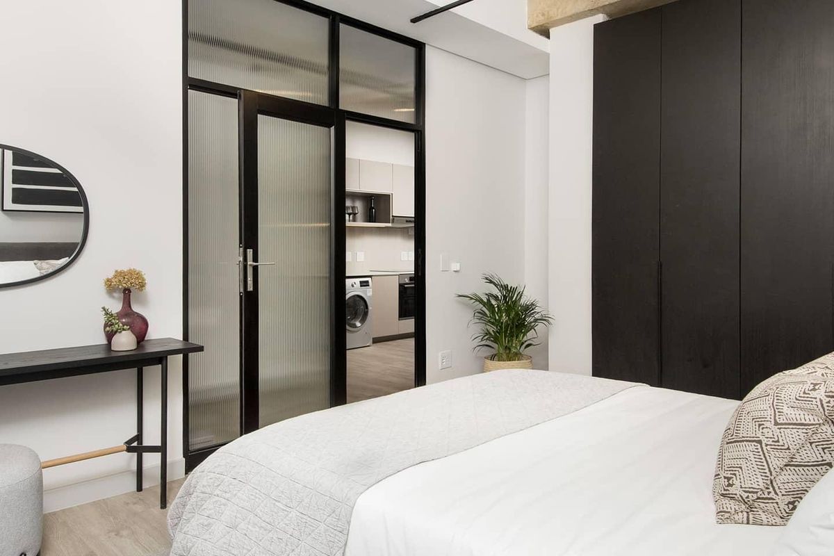 Modern bedroom featuring exposed concrete ceiling, track lighting, and sleek floor-to-ceiling built-in cupboards, with a glass-paneled door leading to the kitchen area.