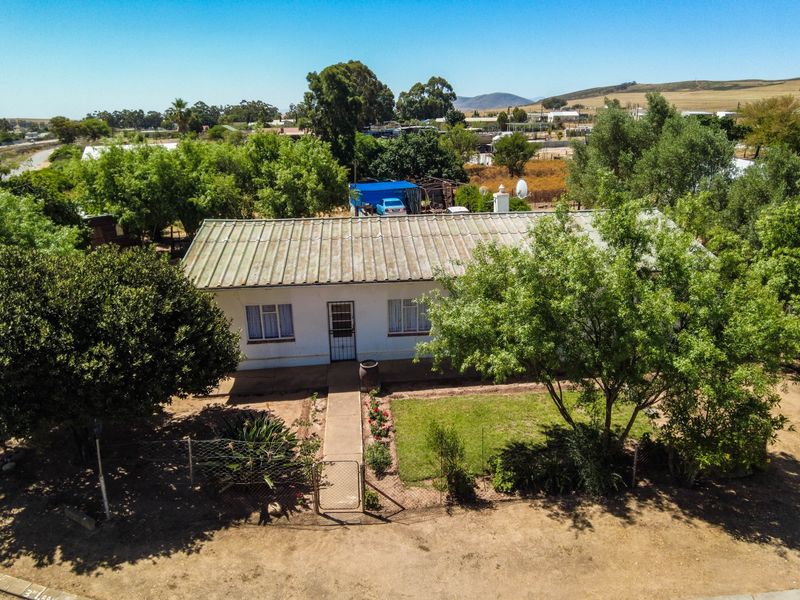 Aerial view of a single-story dwelling with a corrugated metal roof, featuring a front garden, walkway, and surrounding rural landscape.