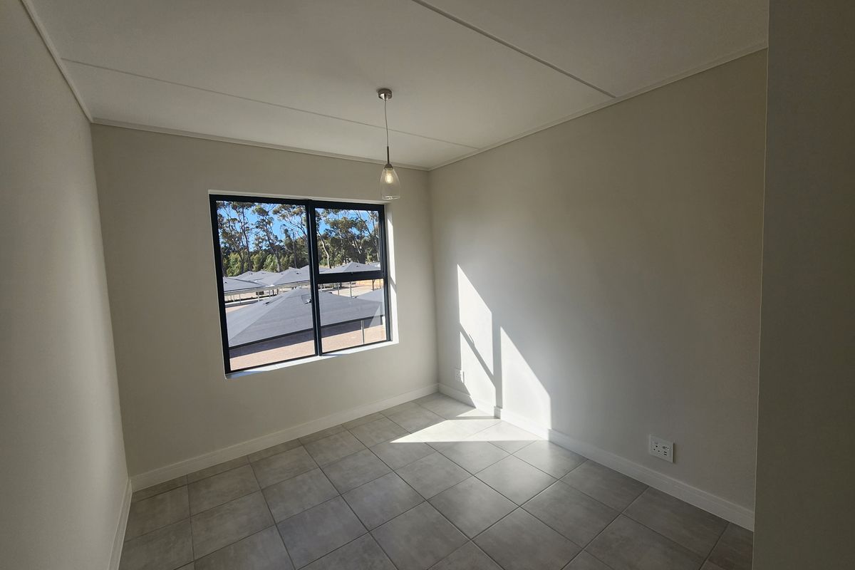A bright, empty bedroom featuring neutral tiled flooring, a modern pendant light, and a large window with views of the surrounding complex.