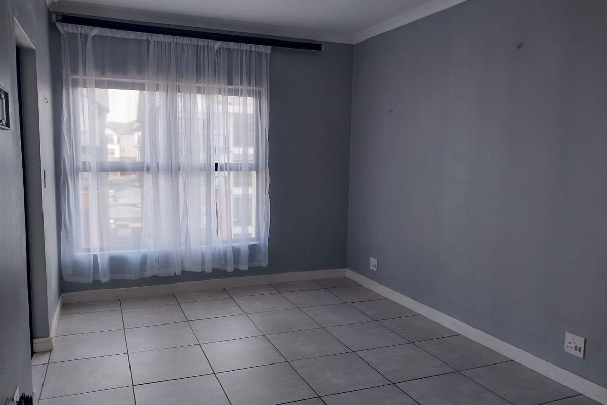 A bright, empty bedroom featuring neutral grey walls, tiled flooring, and a large window with fitted curtain tracks.