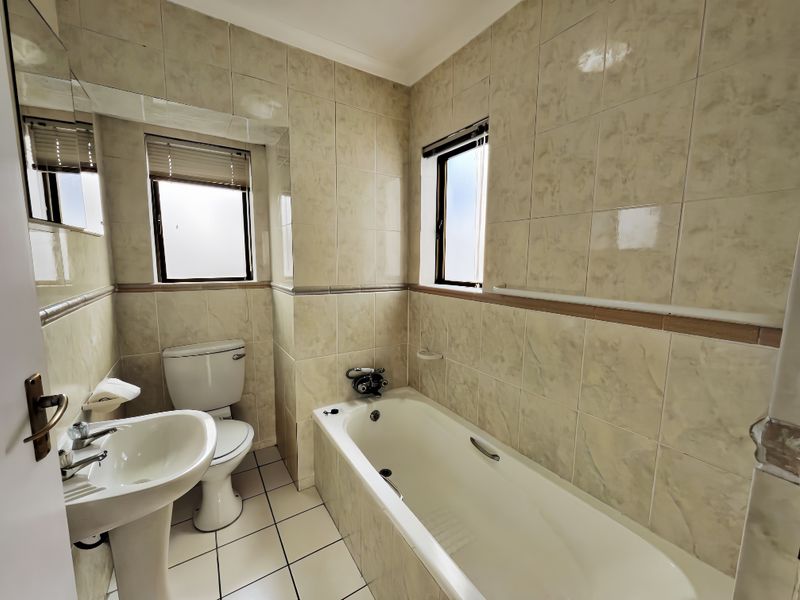 A functional bathroom featuring a pedestal basin, toilet, and bathtub with full-height wall tiling and natural light from two frosted windows.