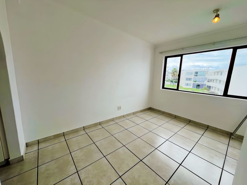 A bright, empty bedroom featuring neutral tiled flooring, a large window with exterior views, and a simple ceiling light fixture.