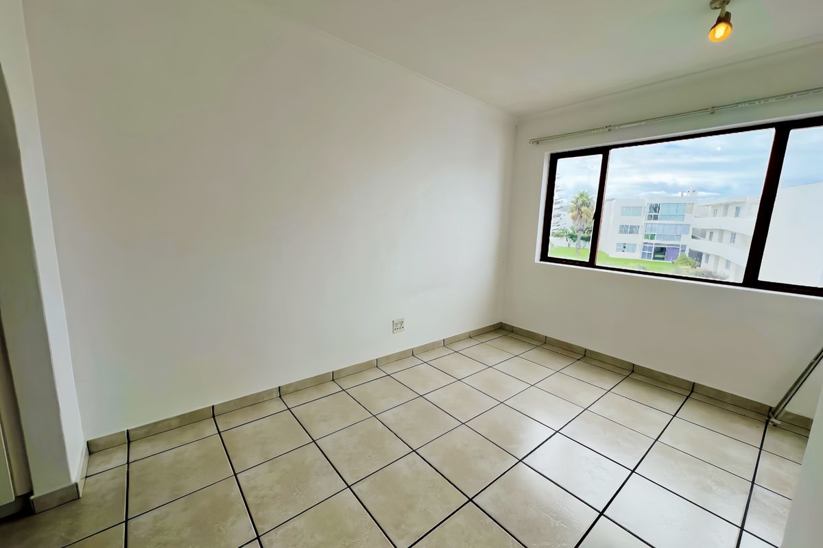 A bright, empty bedroom featuring neutral tiled flooring, a large window with exterior views, and a simple ceiling light fixture.