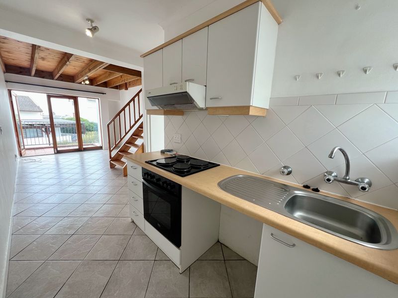 Open-plan kitchen and living area featuring tiled floors, built-in cabinetry, exposed timber ceiling beams, and direct access to a balcony via sliding glass doors.