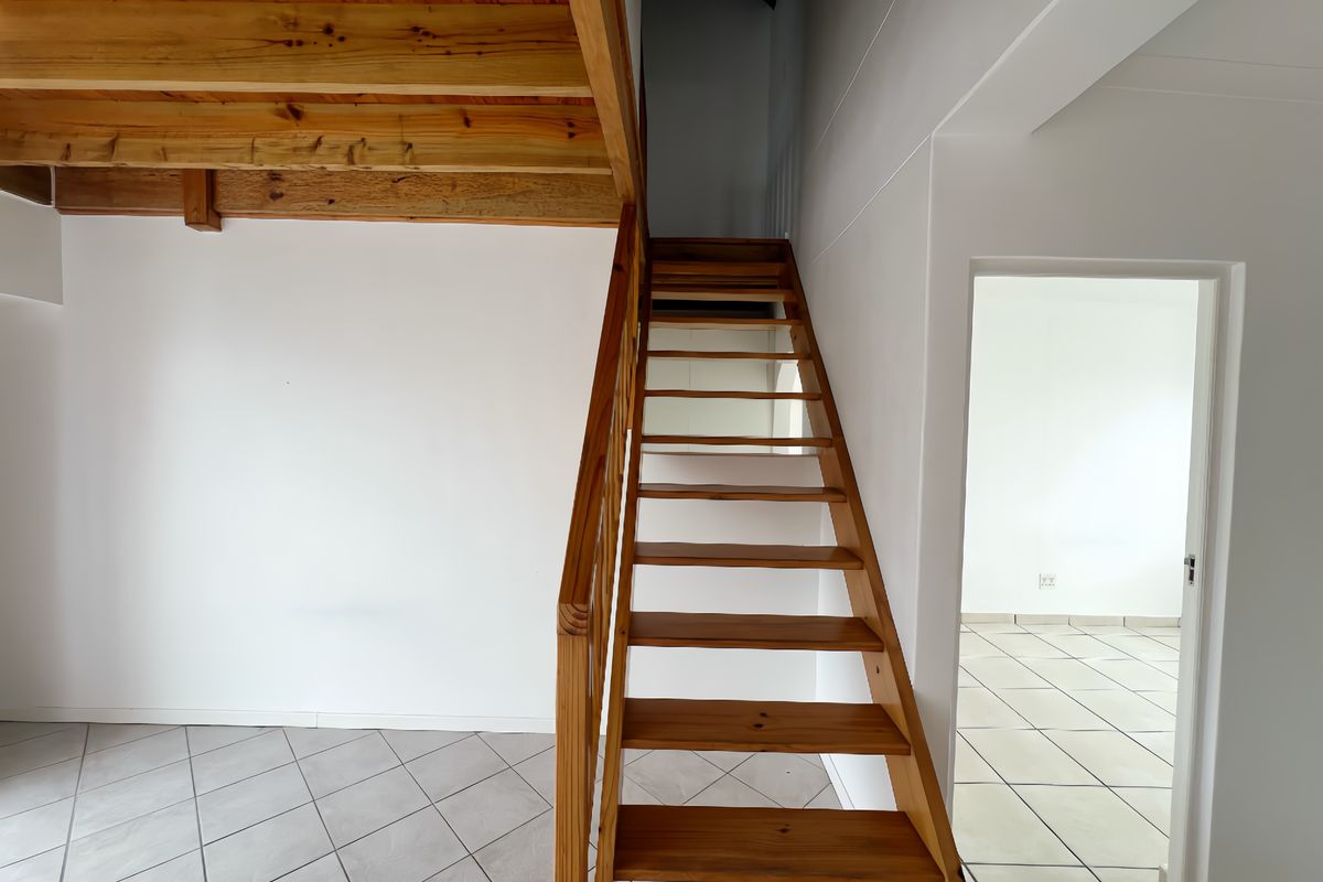An open-plan living area featuring a wooden staircase leading to a mezzanine level, with neutral tiled flooring and clean white walls.