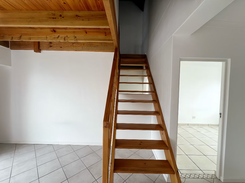An open-plan living area featuring a wooden staircase leading to a mezzanine level, with neutral tiled flooring and clean white walls.