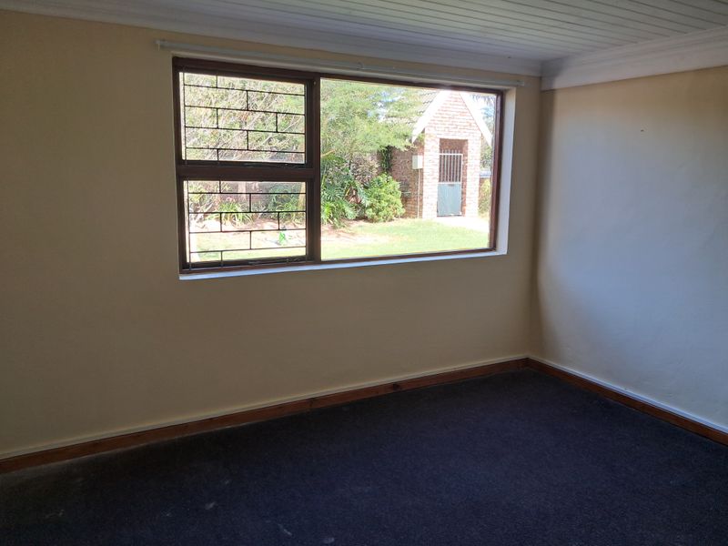 An empty bedroom featuring dark carpeting, timber skirting boards, and a large window with security bars overlooking the garden.