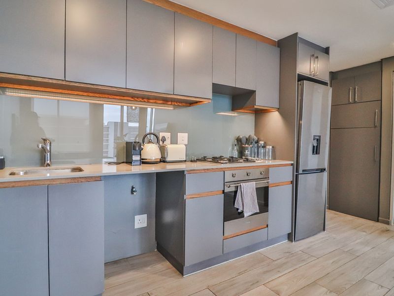 Modern kitchen featuring sleek grey cabinetry, stone countertops, glass splashback, integrated appliances, and high-quality wood-look tile flooring with warm under-cabinet lighting.