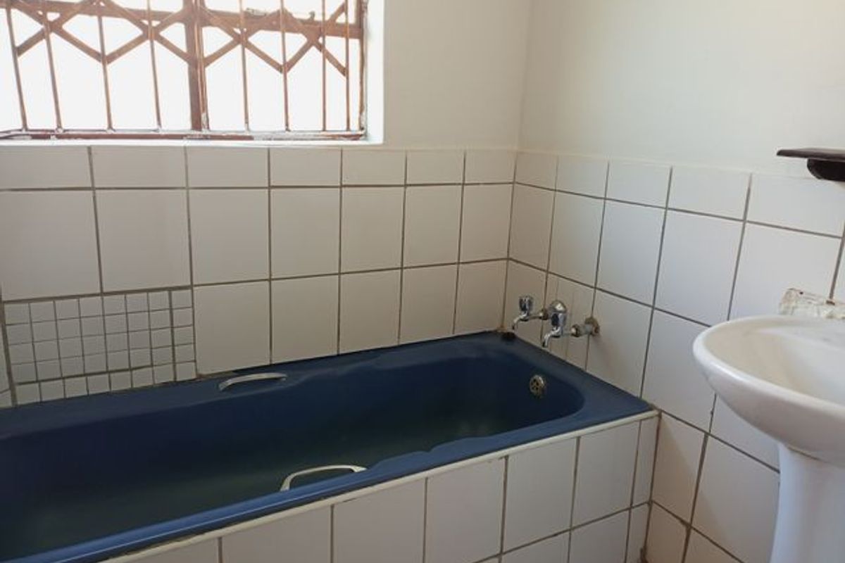 A dated bathroom featuring a blue bathtub, pedestal basin, and white wall tiling with security bars on the window.