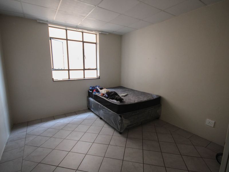 A simple bedroom featuring tiled flooring, a drop ceiling, and a steel frame window, providing a basic functional space.