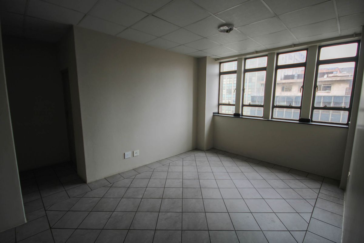 An empty, open-plan living area featuring tiled flooring, a drop ceiling, and large windows providing natural light.