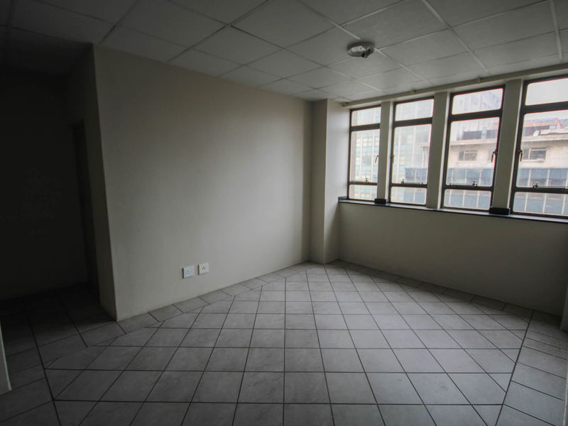 An empty, open-plan living area featuring tiled flooring, a drop ceiling, and large windows providing natural light.