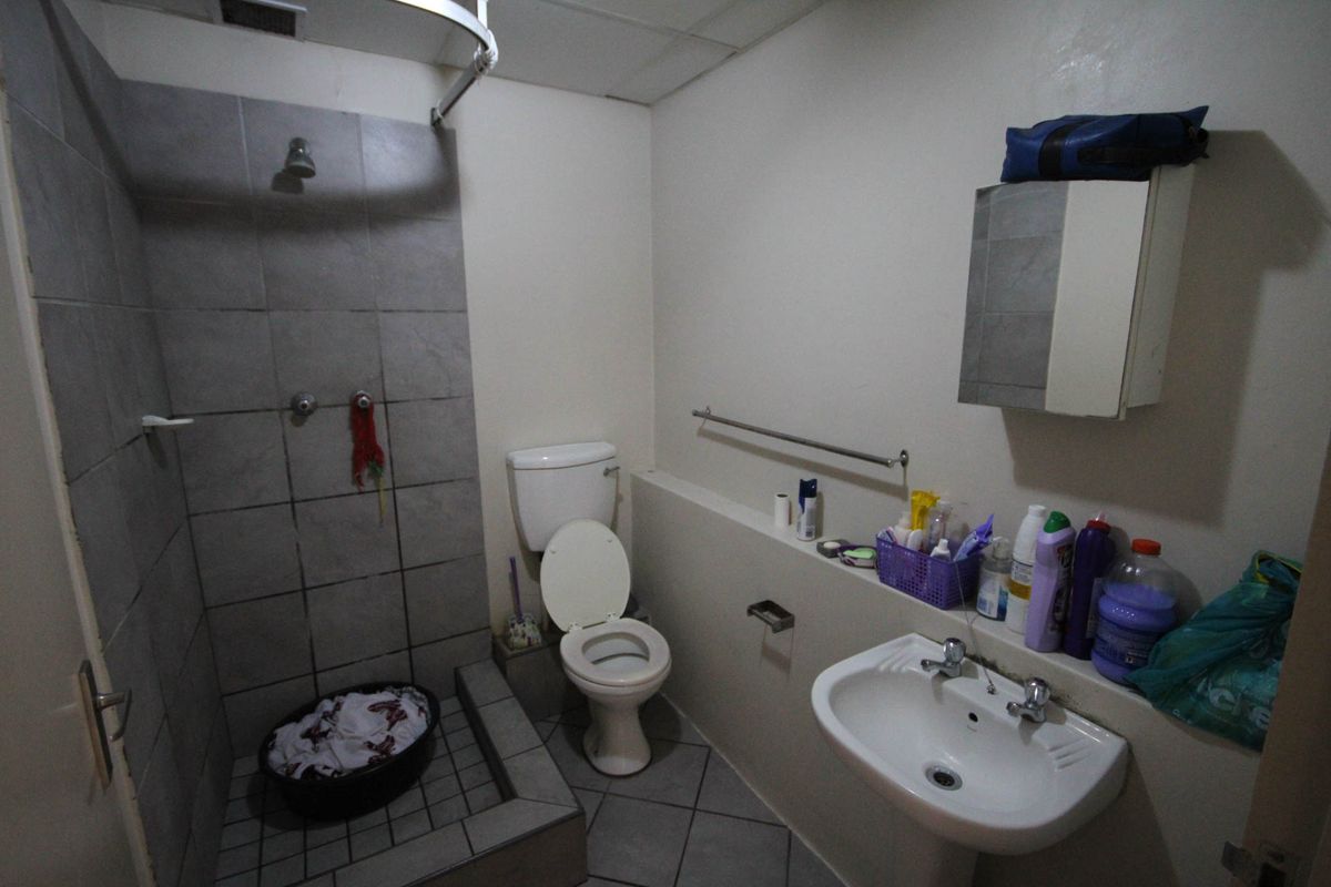 A functional bathroom featuring a tiled shower enclosure, pedestal sink, and toilet, with dated finishes and visible wear on the floor tiles.
