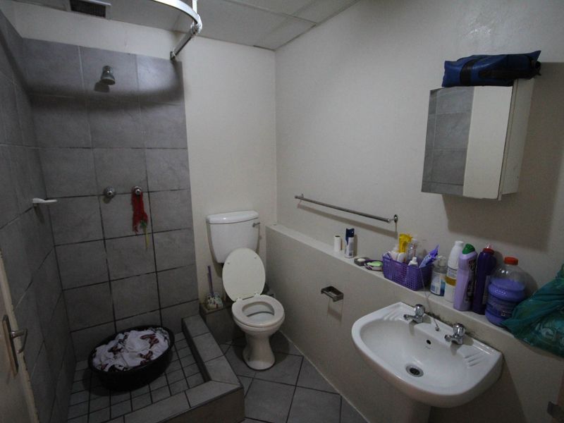 A functional bathroom featuring a tiled shower enclosure, pedestal sink, and toilet, with dated finishes and visible wear on the floor tiles.