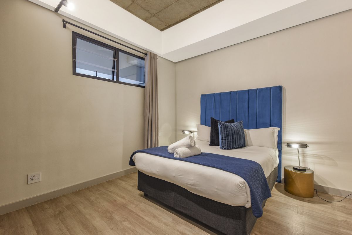 A modern bedroom featuring laminate flooring, an exposed concrete ceiling with cove lighting, and a high-set aluminium window.