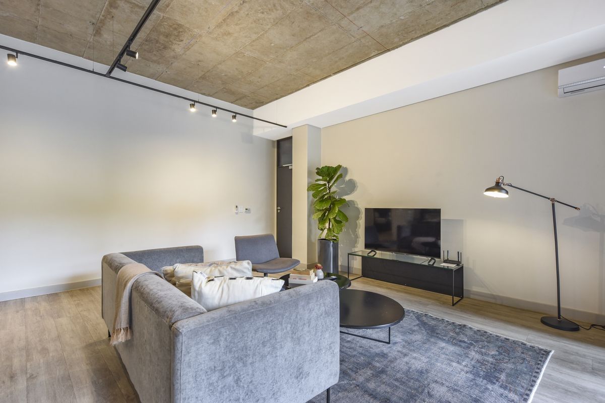 Modern living room featuring an exposed concrete ceiling, track lighting, and a wall-mounted air conditioning unit with durable laminate flooring.