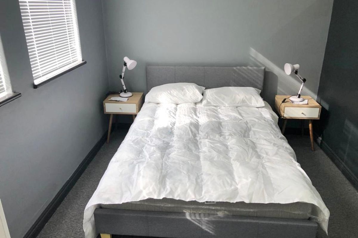A well-lit bedroom featuring neutral grey walls, carpeted flooring, and venetian blinds, providing a clean and comfortable living space.