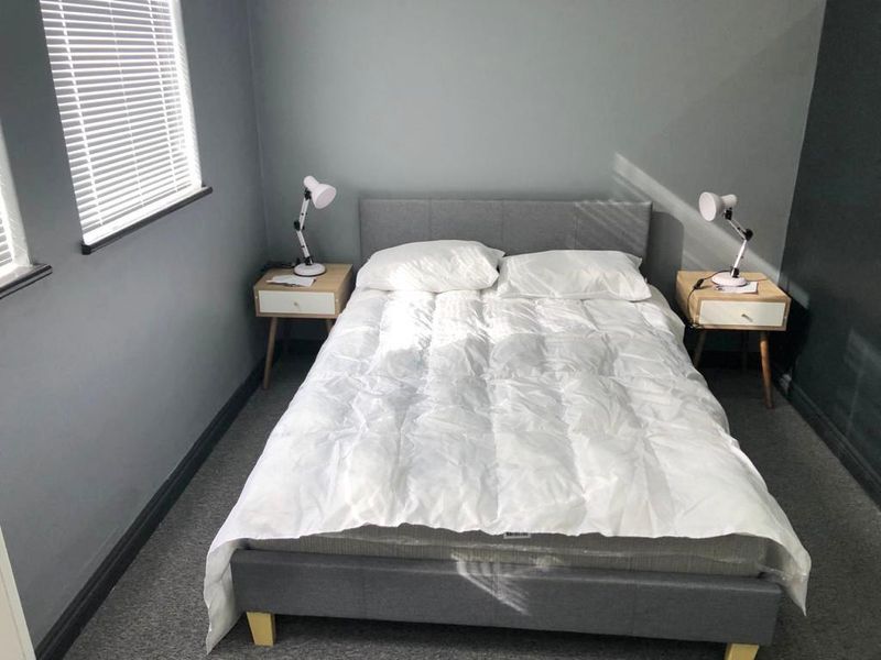 A well-lit bedroom featuring neutral grey walls, carpeted flooring, and venetian blinds, providing a clean and comfortable living space.