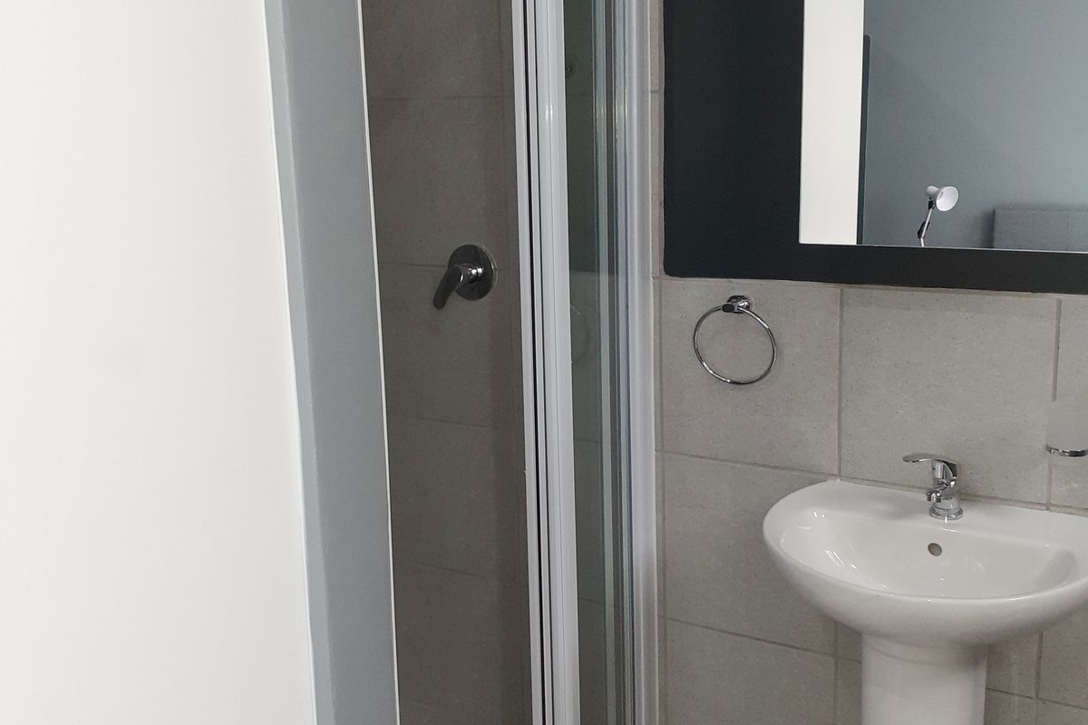 A clean, modern bathroom featuring a pedestal basin, wall-mounted mirror, and a glass-enclosed shower with neutral wall tiling.