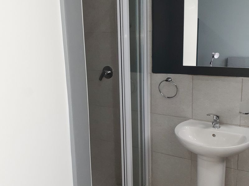 A clean, modern bathroom featuring a pedestal basin, wall-mounted mirror, and a glass-enclosed shower with neutral wall tiling.