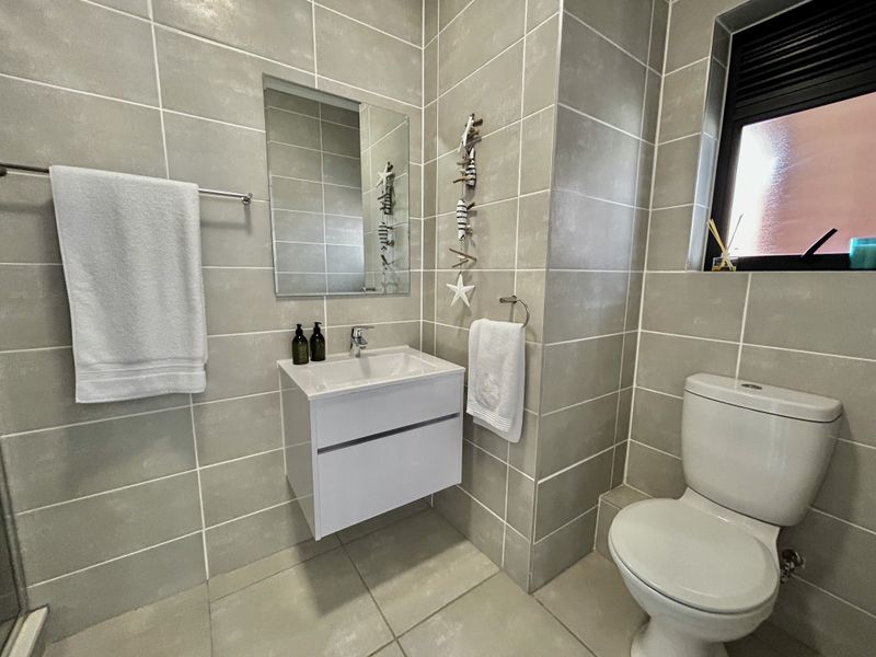 Modern bathroom featuring neutral grey wall and floor tiling, a wall-mounted vanity with basin, and a dual-flush toilet.